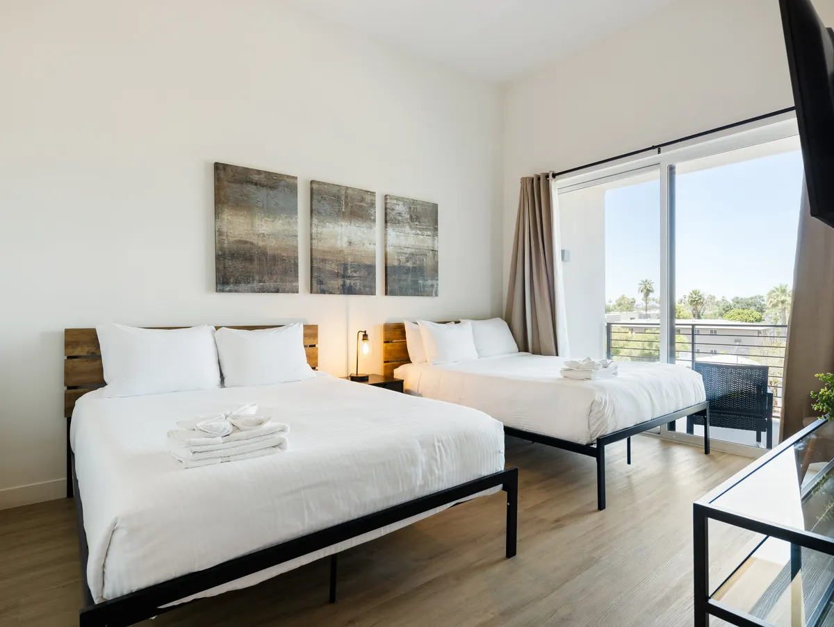 A peaceful bedroom scene with two beds, abstract wall art above the beds, and a balcony visible through large windows, inviting natural light. The view from the balcony and the arrangement of the beds emphasize both comfort and indoor-outdoor connection.