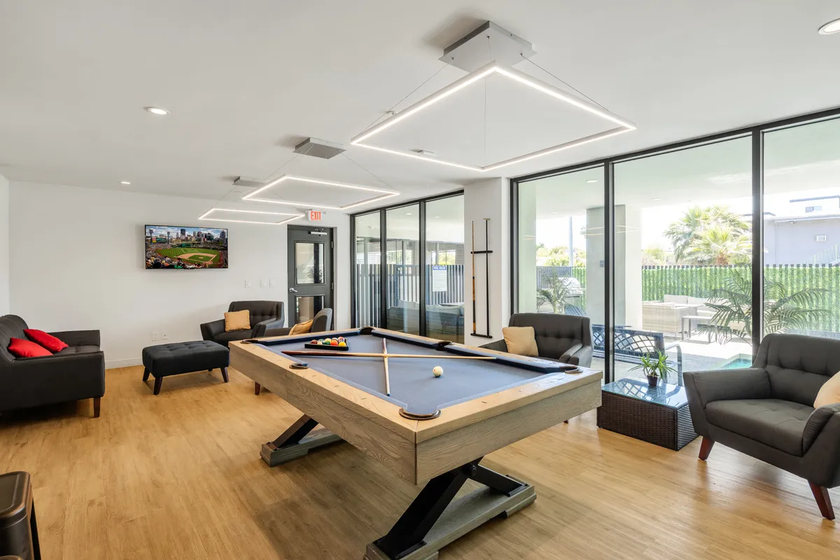 A spacious clubhouse interior with a pool table as the centerpiece, surrounded by plush sofas and a television. Large windows bring in natural light, creating a friendly and inviting atmosphere suitable for social gatherings and recreation.