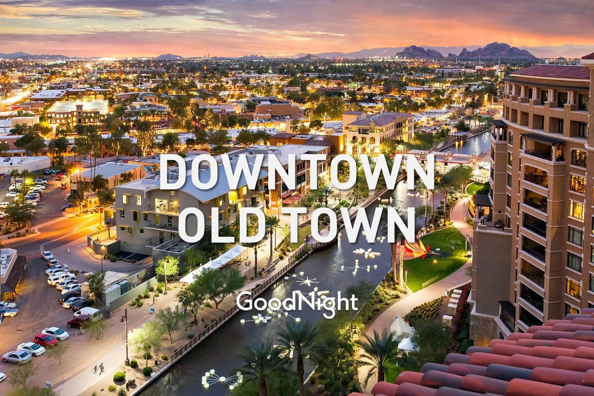 A scenic twilight view of illuminated modern buildings lining a canal, with palm trees and a mountain backdrop. The lively canal area is bustling with people strolling, dining outdoors, and shopping, encapsulating the urban vibrancy of Scottsdale’s city center.