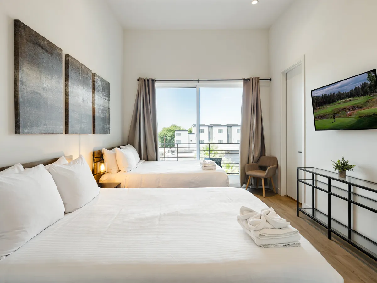 A bright, serene bedroom featuring two beds with crisp white linens, a large window allowing in natural light, decorative artwork on the wall, a wall-mounted Smart TV, and minimalist furniture. The spaciousness and setup suggest comfort and a restful atmosphere ideal for rejuvenation.