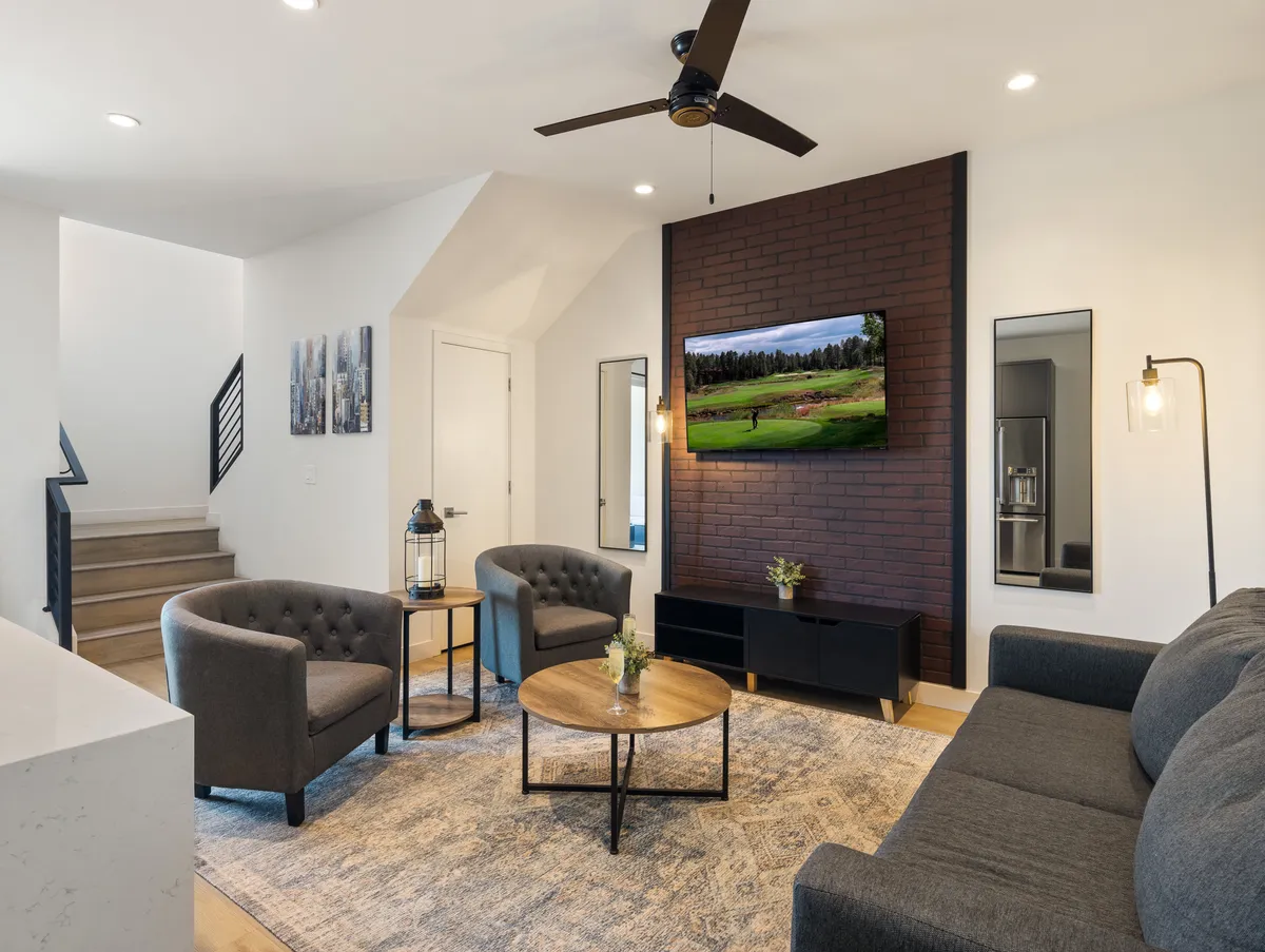 The image showcases a modern, sunlit living room with a striking brick accent wall, a large TV displaying a scenic view, plush and stylish seating, and a chic coffee table—creating a perfect space for both relaxation and socializing. The inviting setup suggests comfort, warmth, and an ideal spot for gathering with friends or unwinding after a day in lively Old Town Scottsdale.