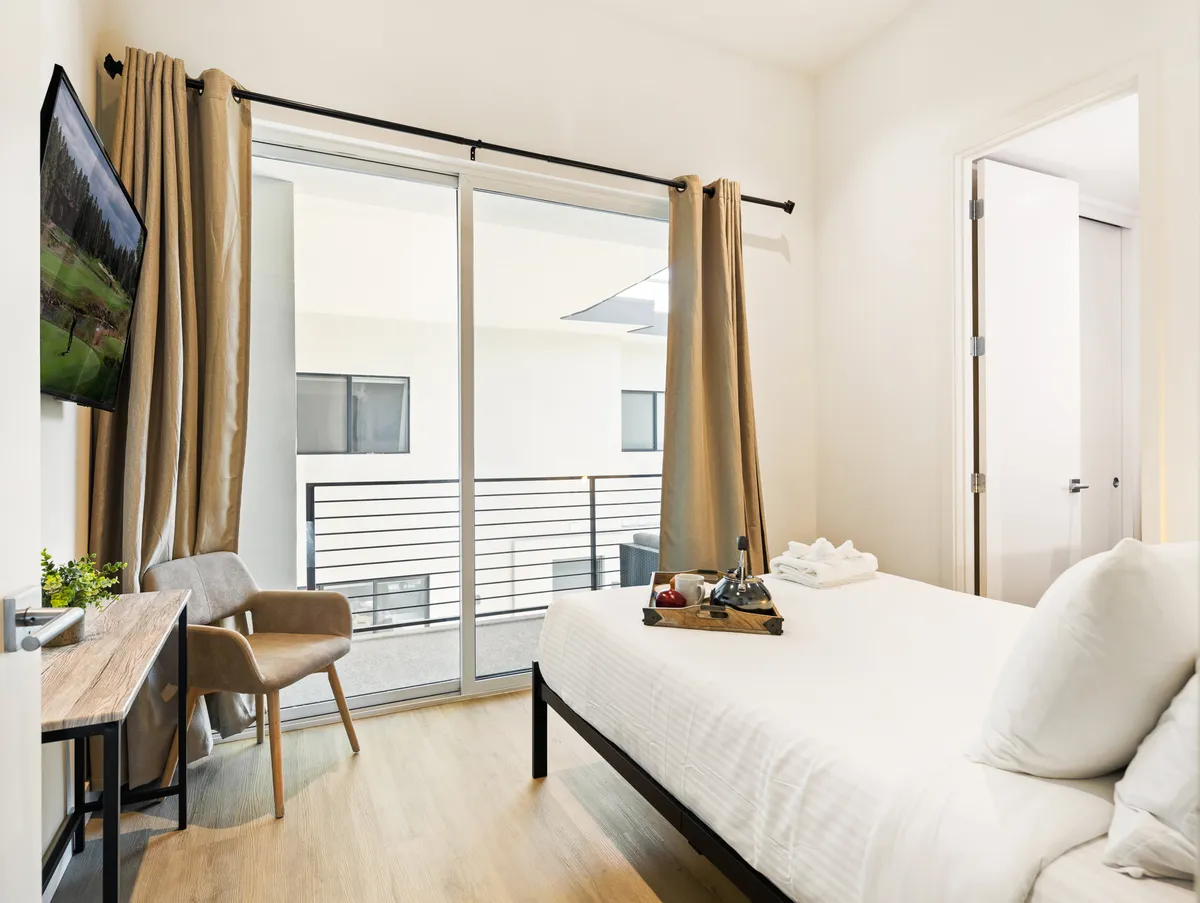 A cozy, modern bedroom with a Full-size bed, TV mounted on the wall, sliding glass door leading to a balcony, a small wooden desk with a chair, and a decorative plant. The space is inviting and designed for relaxation or a quiet moment to work or dine.
