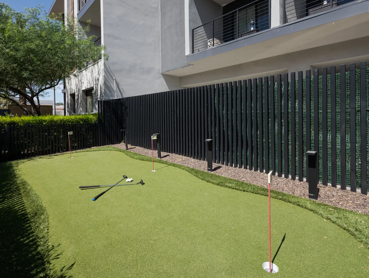 A well-maintained putting green set against modern architecture and fenced surroundings. Golf clubs rest on the green, suggesting active use and moments of leisure, relaxation, and friendly competition among residents.